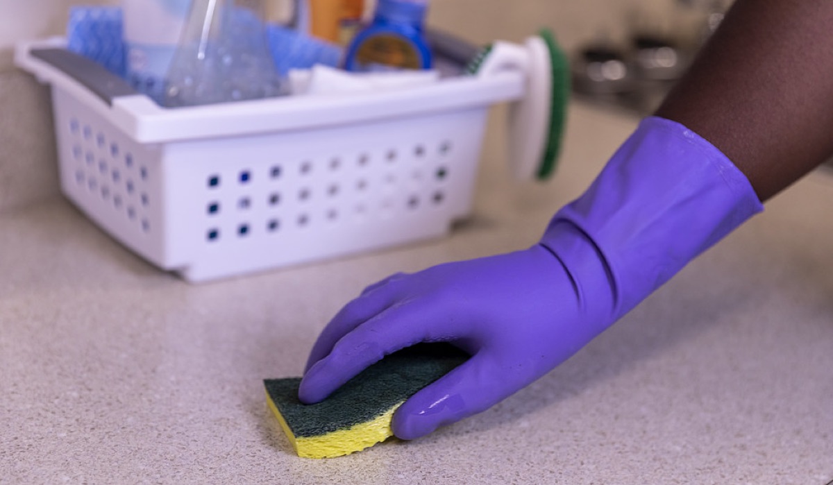 A gloved hand scrubbing a counter with a sponge, a cleaning caddy nearby